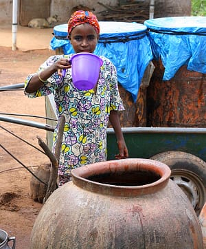 One of Abdullah’s daughters takes a drink of water from a clay water storage jar. The donkey cart and drums used for transporting water to the house stand behind her.